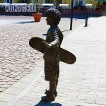 A bronze metal art sculpture of a boy holding a skateboard outdoors